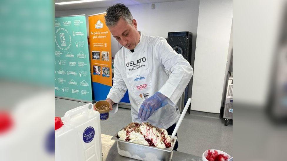 Daniele Panunzio competing in a gelato-making contest, wearing blue plastic gloves and a white chef's jacket with a contestant bib pinned to the front. He is sprinkling a tub of caramelised peanuts over a metal tray of white ice cream, drizzled with raspberry sauce. He is standing in grey commercial kitchen behind a metal counter. 