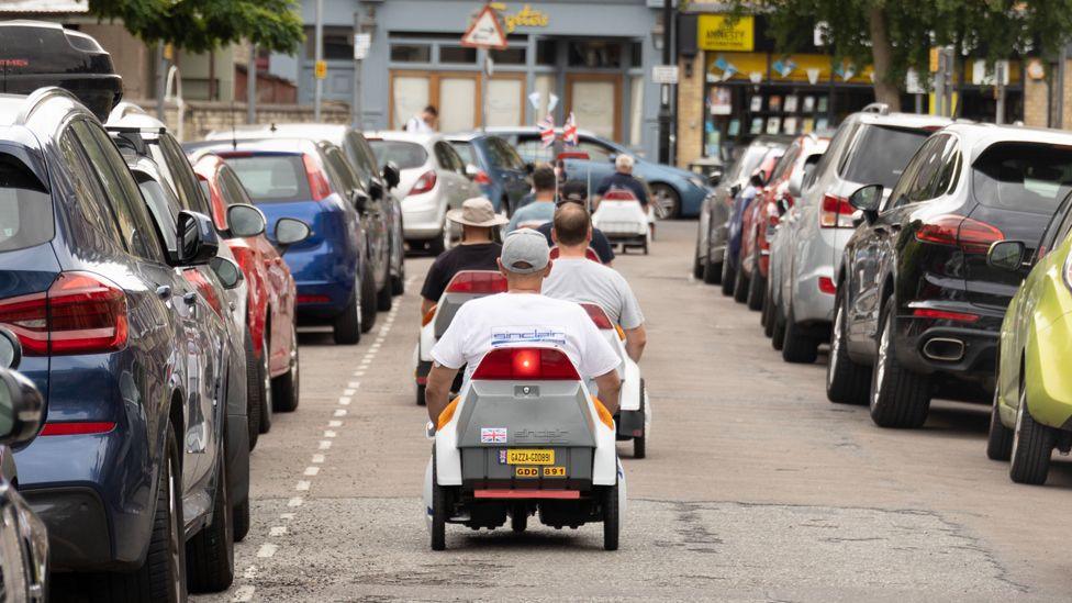 About six Sinclair C5 vehicles are seen from behind in a row as they travel down a residential street. There are cars parked on both sides of the street and some shops in the background