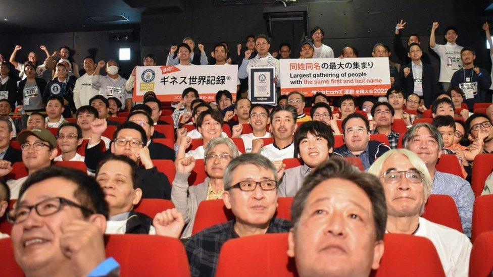 Group of men in a theatre, holding a poster reading "largest gathering of people with the same first and last name"