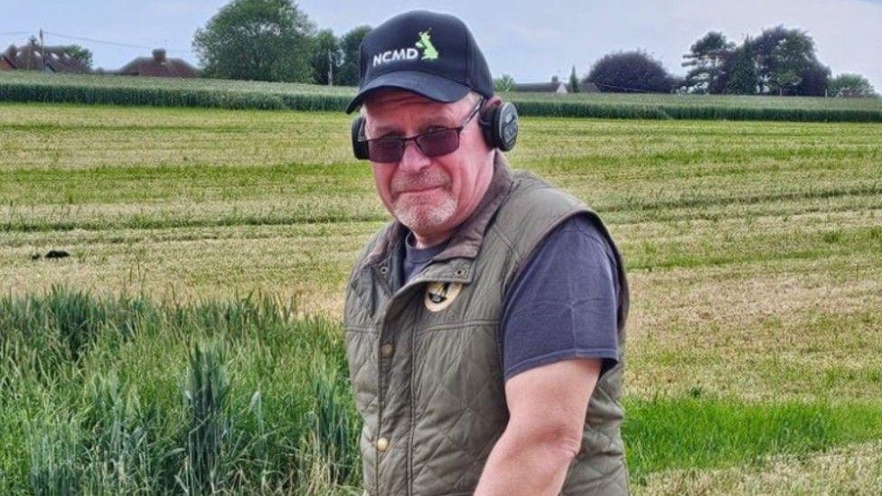 Steve Hickman standing in a green field. He is wearing a blue T-shirt with a khaki gilet over the top. He is wearing a pair of sunglasses and a baseball cap and is looking directly at the camera.