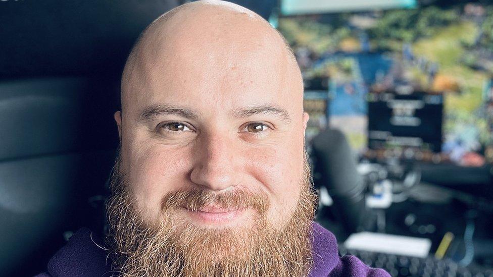 A man with a fairly long, grey beard and bald head smiles warmly. He's sitting in front of a large computer monitor displaying a strategy game. It's blurred but we can just make out trees, grass and lakes and units dotted around the map. The outline of a microphone and keyboard are just visible.