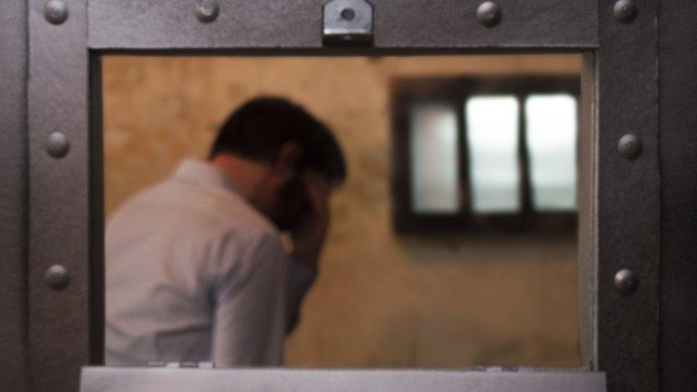 A man stands in a prison cell by a barred window, his head in his hand.