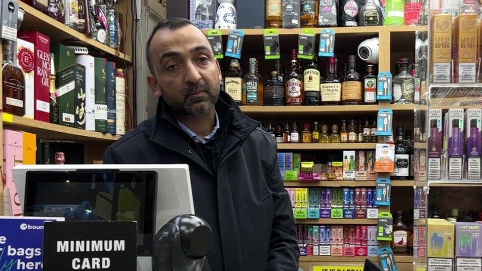 Image of Chandra Goyal wearing a dark blue jacket behind the cash desk, in front of shelves of bottles