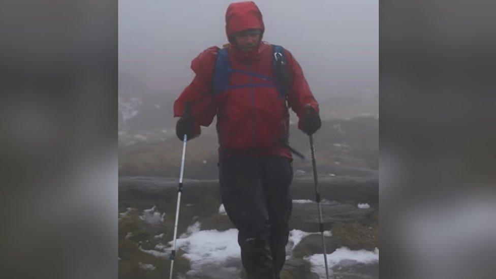 Ian Evans participating in the Winter Spine Challenge in January 2023. He is wearing a red coat and walking with hiking sticks. He is along the Pennine Way and a small amount of snow can be seen on the ground.