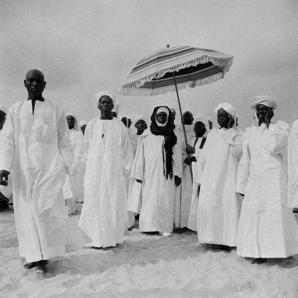 Men dressed in white boubous and turbans walk together. One of them is sheltered from the sun by another man who holds a parasol over his head.