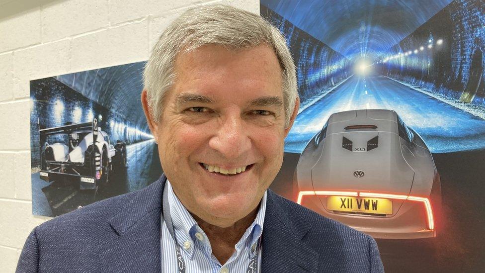 Smiling man with silver-coloured hair stands in front of posters showing cars