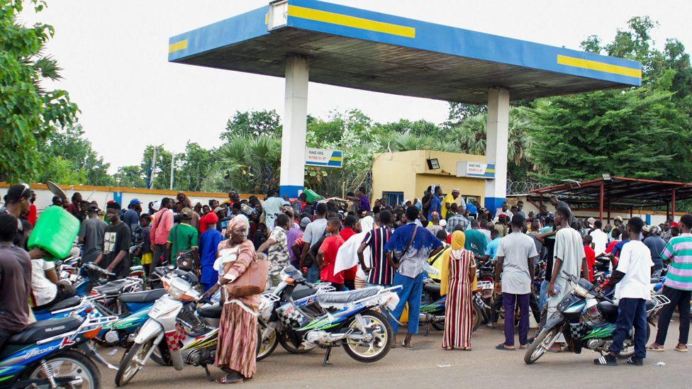 A crowd of people with jerry cans and motorbikes crowd around a garage waiting for fuel in Bamako, Mali - October 2025.