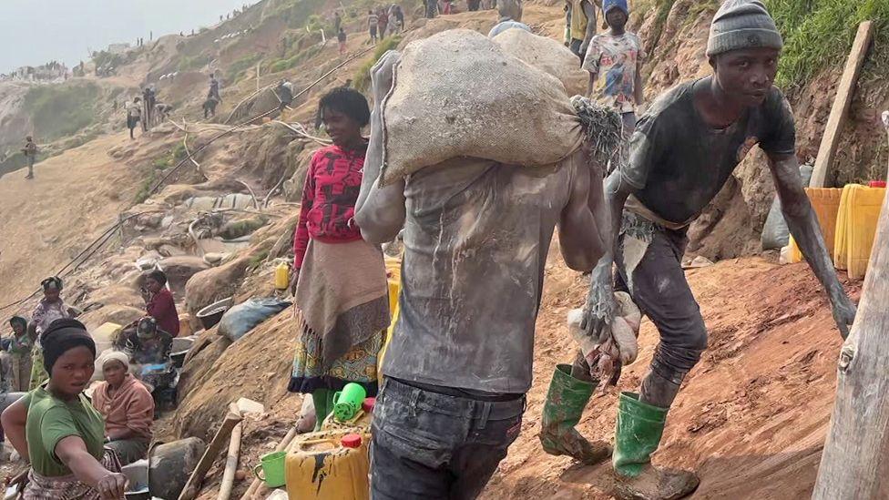 Muddy workers are seen on a slope at Rubaya mine in DR Congo. One in green wellingtons, black jeans and T-shirt looks at the camera, another in a similar outfit is seen from the back as he walks with a sack on his shoulders. A group of women are seen a little further down the slope near some big basins and jerry cans.