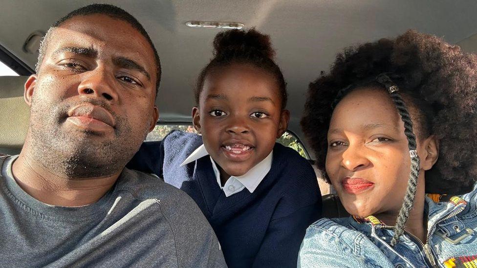 Sipho (L) and Sibahle Mbasana pose for the camera in their car with their daughter Buhle, in navy jumper and white shirt school uniform, smiling between them as she leans in from the back seat.