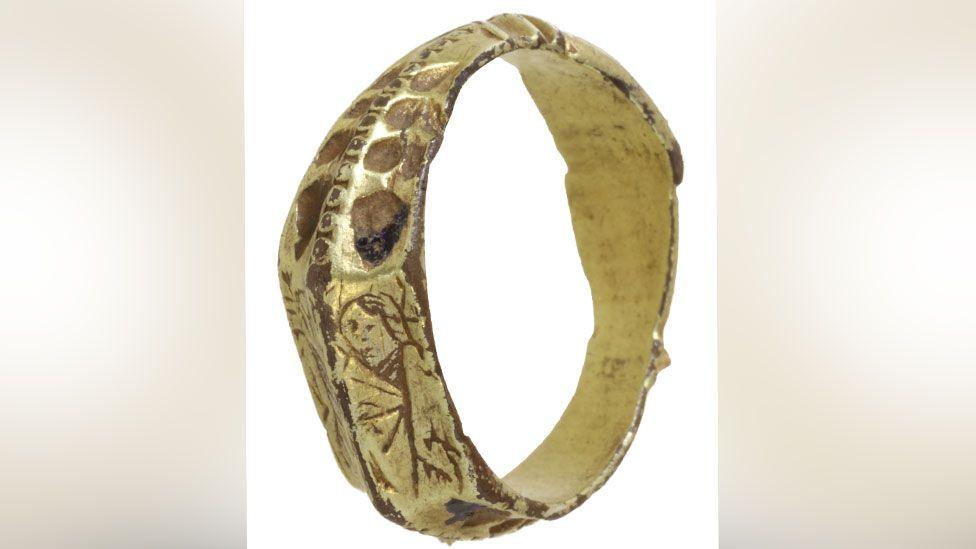 A late medieval gilded silver ring resting on its side so its plain interior can be seen. The exterior is highly decorated and the side facing the viewer shows a crude image of a woman holding a palm. 