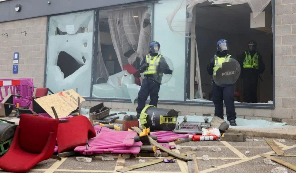 Three police officers stand by a smashed glass window next to a pile of debris, including chairs, barriers and bits of wood.