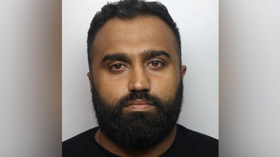 Usman Hanif in his police custody photo. He has a black t-shirt. He has a thick black beard and short black hair with a quiff.