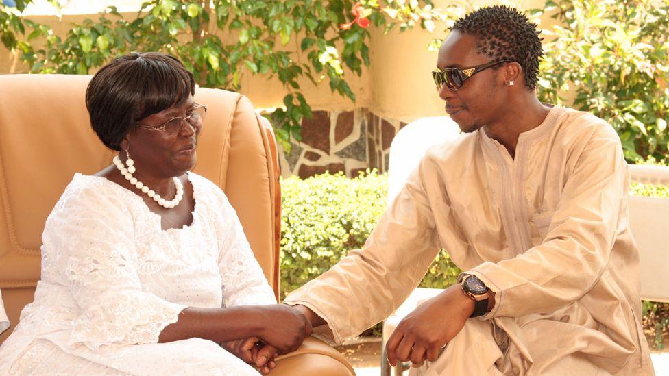 Patience Wilberforce dressed in a white outfit and wearing a white-beaded necklace and earrings sits on a large chair on a veranda holding the hand of her son Mark who sits on a garden chair next to her to the right. He is wearing sunglasses and a light brown kaftan. A courtyard garden can be seen behind them.