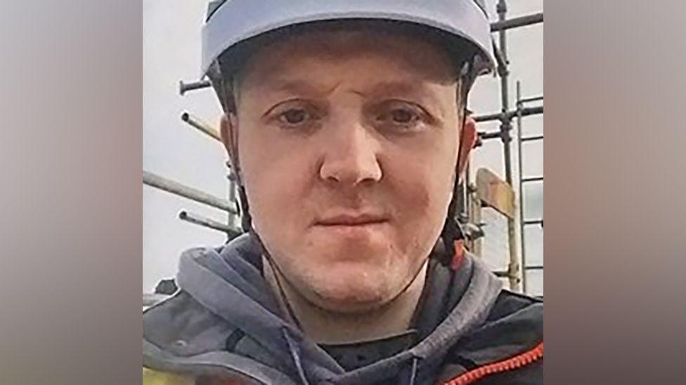 A man looking into the camera while wearing a light blue or grey hard hat, seemingly on a building site. 