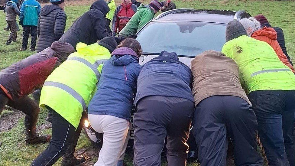 A group of eight people wearing waterproofs pushing a car in a muddy field.