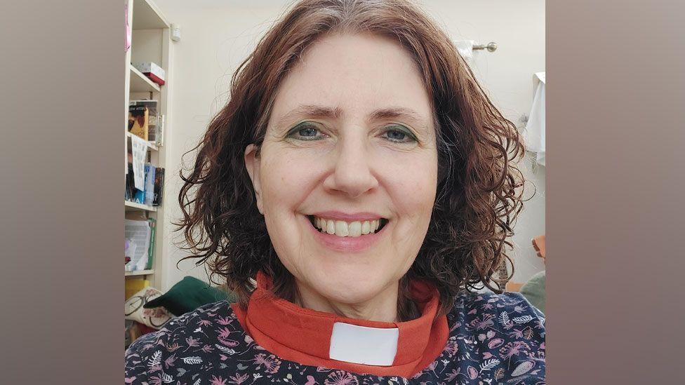 The Reverend Ruth Clay, who has dark, curling chin-length hair and is smiling. She is wearing a red top with a dog collar and over it a dark blue top with a pattern of small leaves