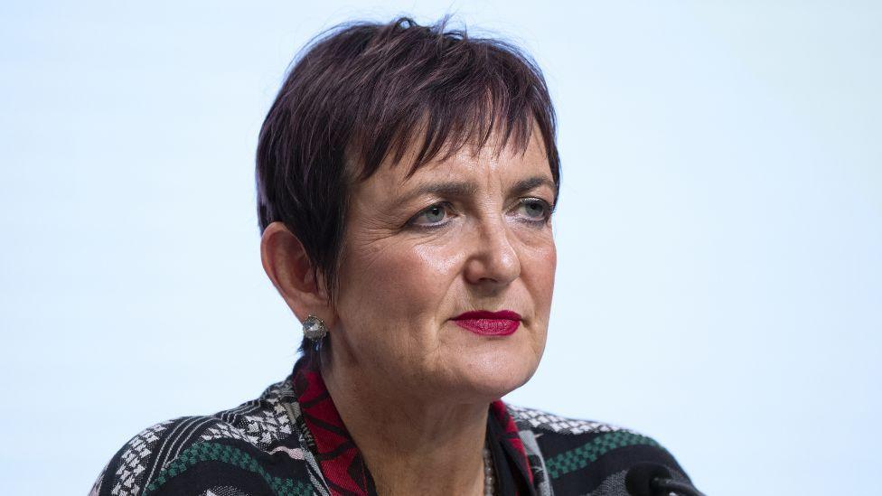 Angela Constance, with dark brown hair, sits in front a light blue background 