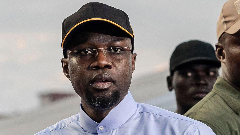 A head and shoulders shot of Ousmane Sonko wearing glasses and in a black cap and buttoned-up blue shirt.