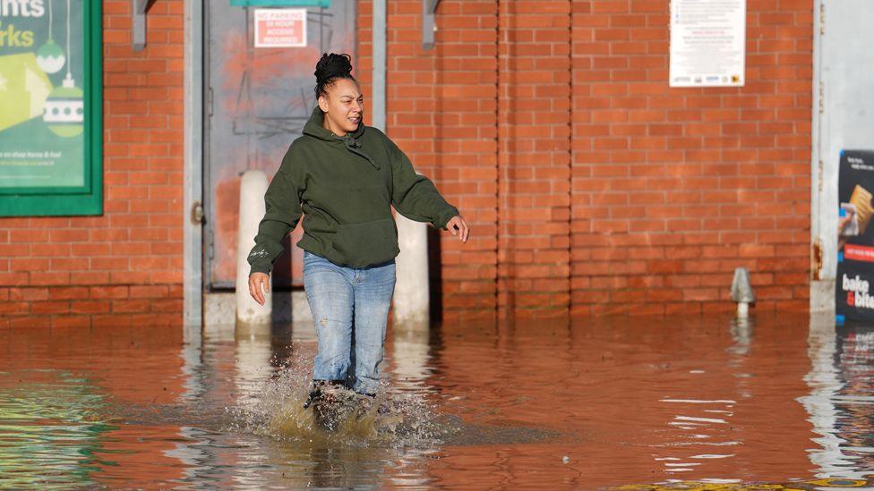 A woman dressed in a hoodie and jeans walks through floodwater outside a building at the Billing Aquadrome in Northamptonshire.