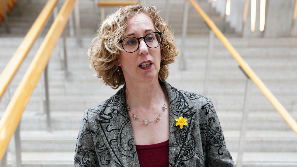 Lorna Slater, who has blonde curly hair and glasses, photographed in a medium close-up shot in front of steps inside the Scottish Parliament. She is wearing a grey patterned jacket and red top.