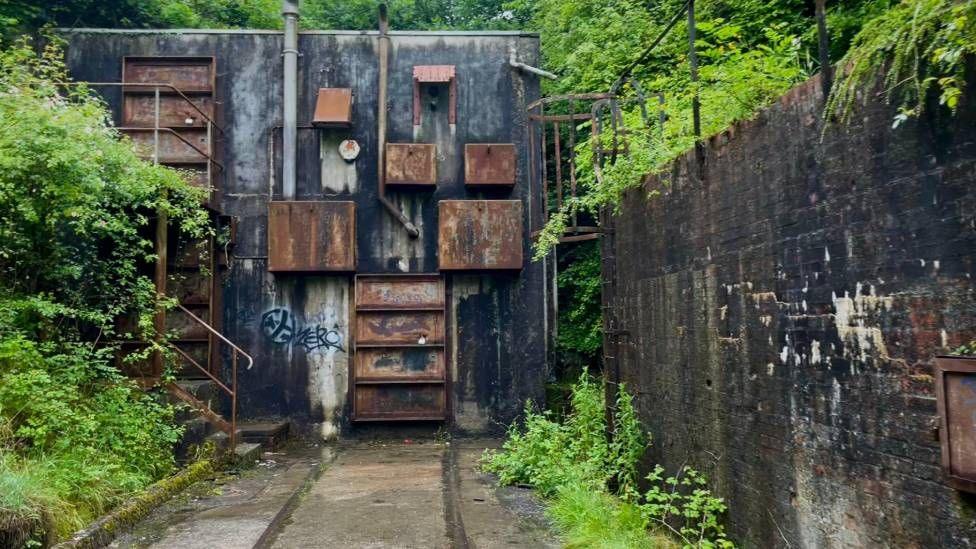 The exterior of a cement building, there are rusty boxes and doors on the front. there is a rail track going all the way down the lane and into the building. There is a storm drain on the left of the tracks and there is overgrowth on all sides.