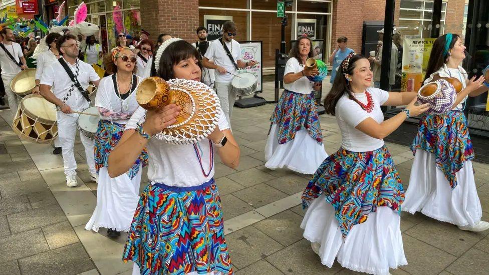 A group of people in colourful costumes, in white and and blues, reds and greens, taking part in a carnival parade in a city centre. They are holding musical instruments.