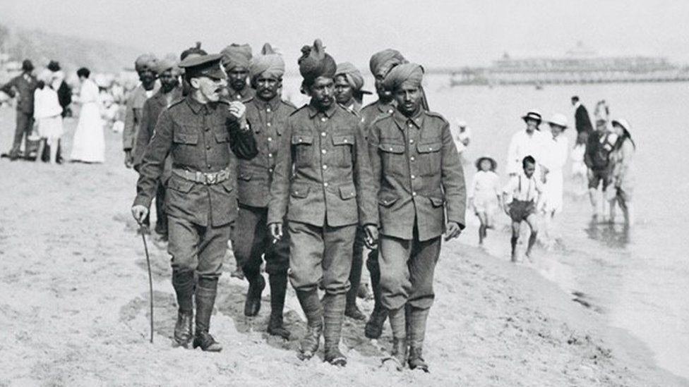 A group of Indian soldiers in uniform walking across beach with pier in background and people padding in the sea