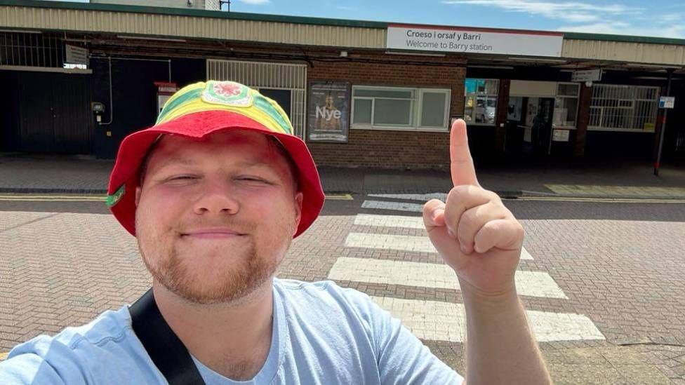 A man in a red, yellow and green bucket hat and light blue t shirt stood in front of Barry train station