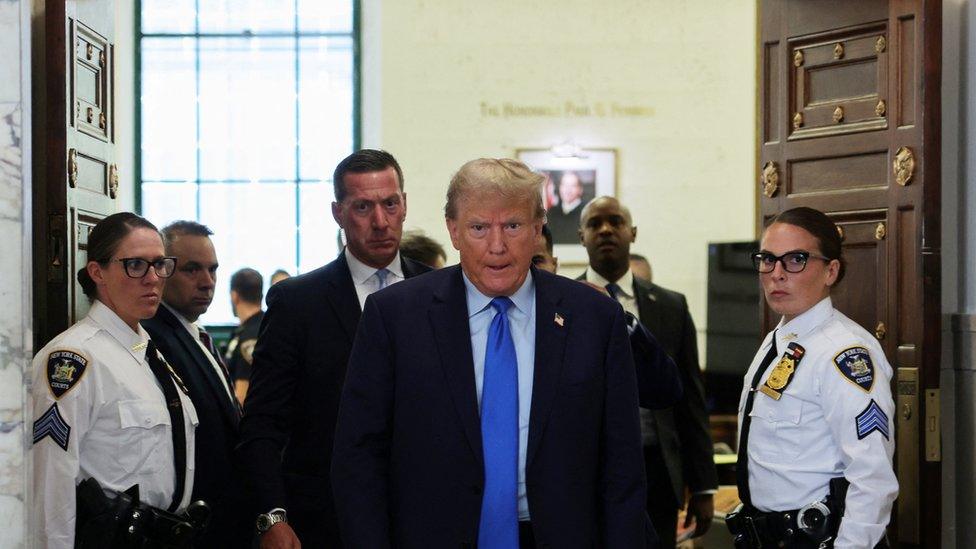 Trump in blue tie and suit, between two security guards and courtroom doors.