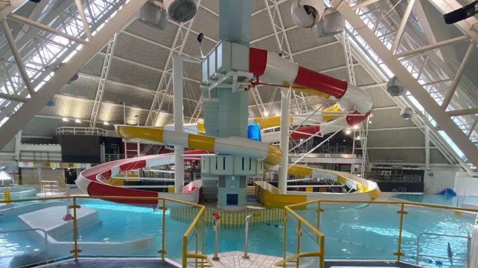 The interior of a white-walled leisure pool, showing lines of windows in the roof, a circular pool, and a chute in the centre. There are spectator seats behind.
