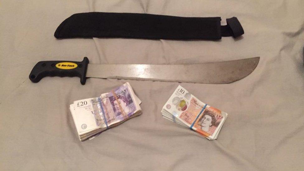 A machete lying on a bed with two wads of bank notes - one is £10 and the other wad contains £20 notes