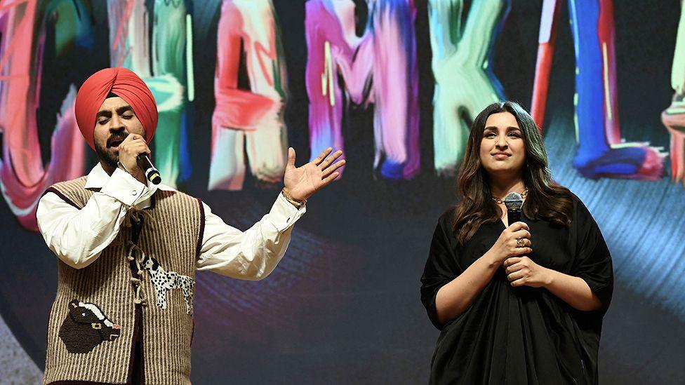 Diljit Dosanjh and Parineeti Chopra at a movie premiere. Diljit is wearing a brown striped vest top with a white shirt and singing into a black mic, while Parineeti has a black outfit and is looking outwards.