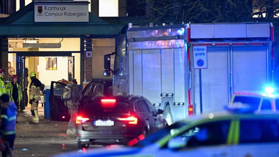 Emergency first responders gather around the Campus Risbergska at night time. Some are wearing firefighters uniforms, while others are wearing military gear.