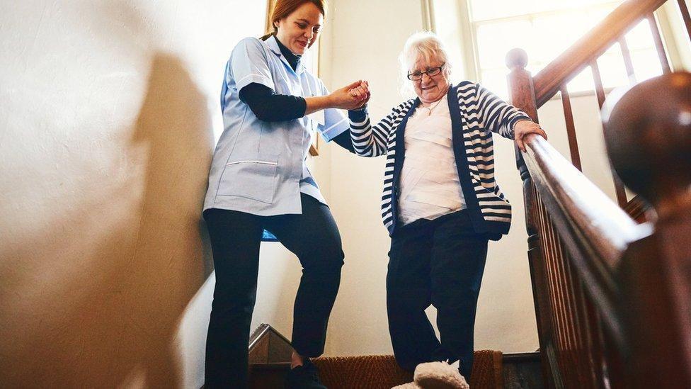 A female care worker in a blue shirt helps an old lady in a striped cardigan walk down stairs by holing her hand