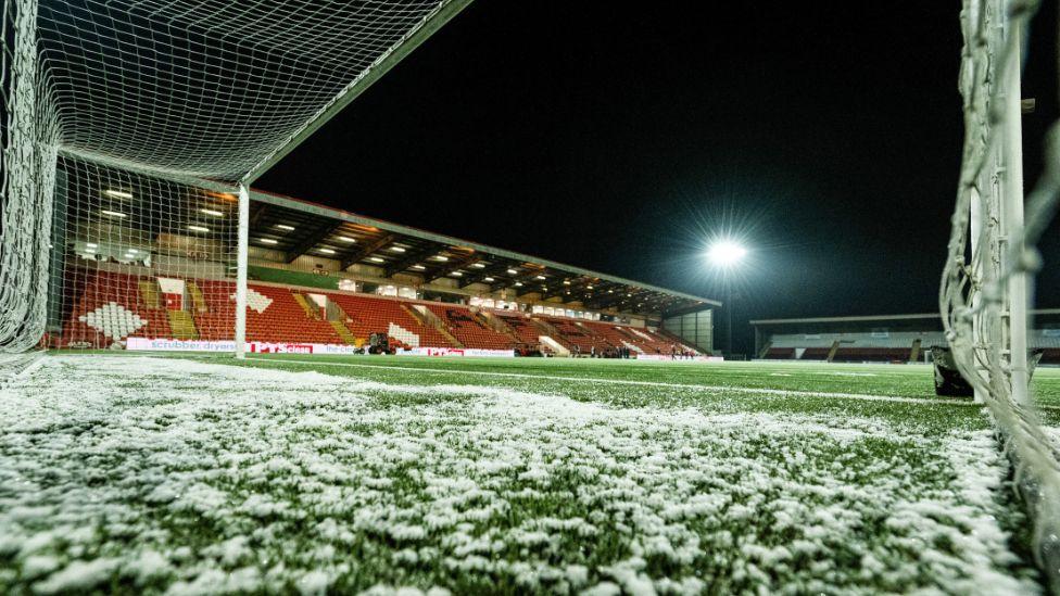 A picture taken from inside a goalmouth of a football stadium. It shows snow lying on the pitch with a red stand in the background. 