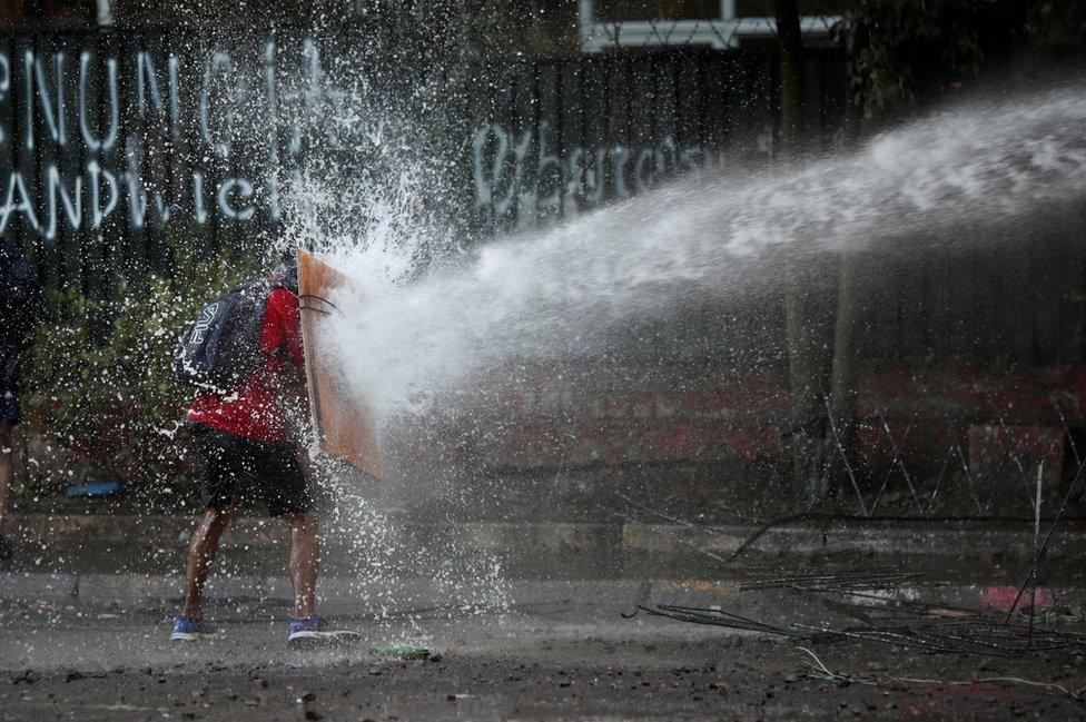 A demonstrator holds a makeshift shield to cover himself from a water cannon aimed by riot police during a protest against Chile's government in Santiago