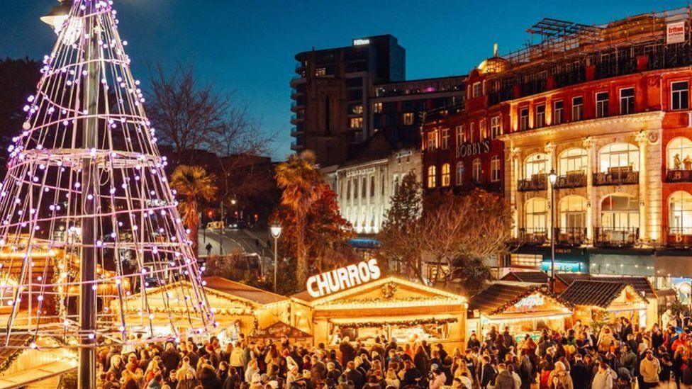 Crowds around lit-up market stalls with a large wire Christmas tree and buildings around the edge.