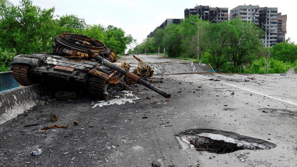 A destroyed tank is pictured in Mariupol - amid Russian military action in Ukraine - with damaged buildings in the background and a large shell hole in the road in the foreground.