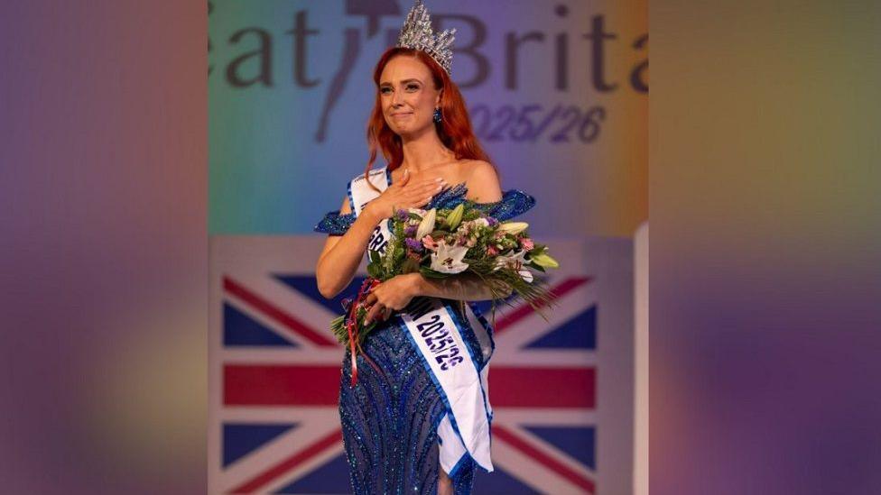Alice Cutler wearing a dark blue sparkly dress, a sash with the words: "Miss Great Britain" and holding a bunch of flowers and a crown in her hair. She has red hair and is stood on stage after winning.