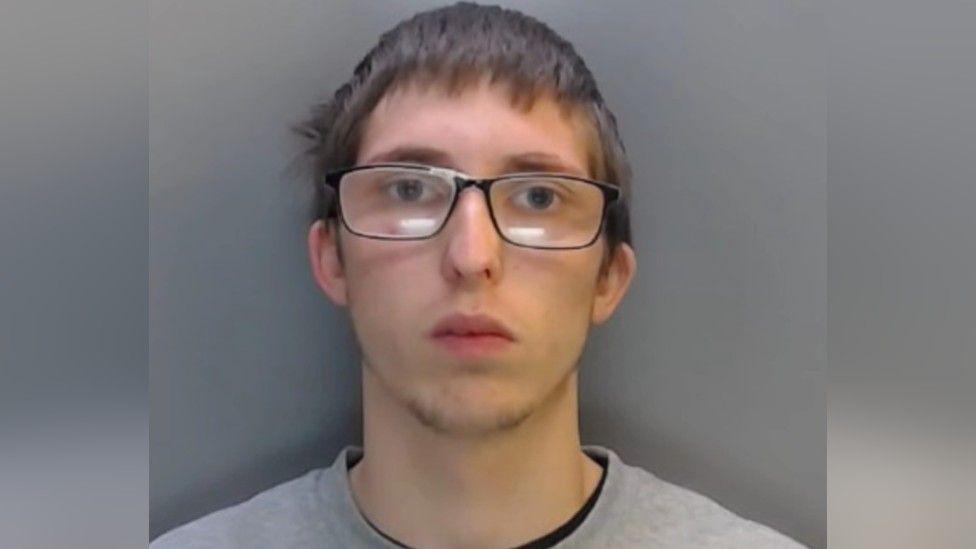 Police mugshot of Aidn Cowen. He has short, unkempt brown hair, thin facial hair at the bottom of his chin and he is wearing glasses. 