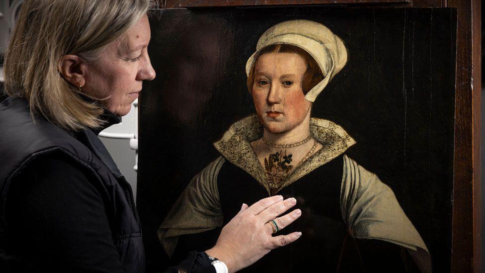 A head and torso portrait of a Tudor woman believed to be Lady Jane Grey. She is wearing a white linen cap, a white lacework open collar, a gold necklace a black dress and a white scarf over its shoulders.  Rachel Turnbull is on the left and is side on, dressed in black with light brown shoulder-length hair examining the portrait. Her right hand is lifted towards the painting.