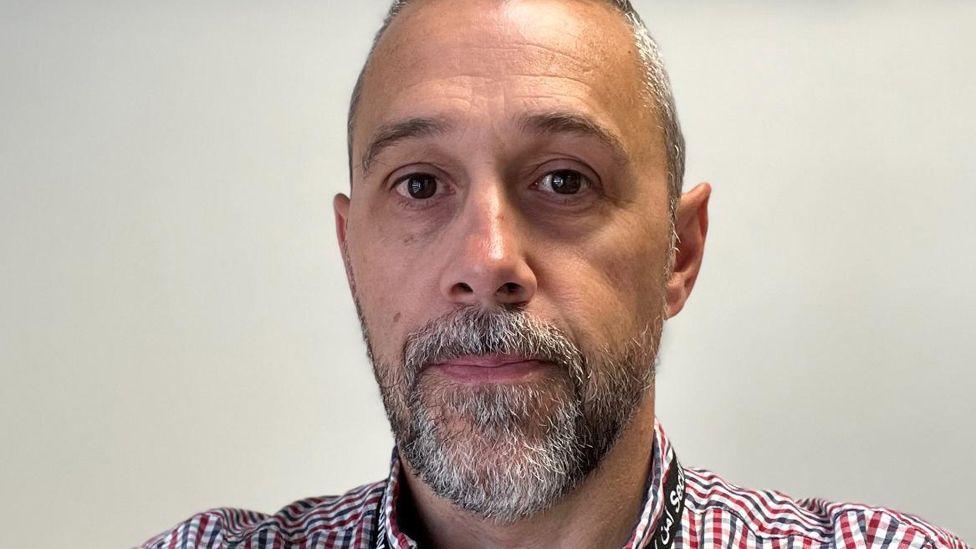 A close-up picture of a man with a grey and black beard staring into the camera wearing a checked shirt