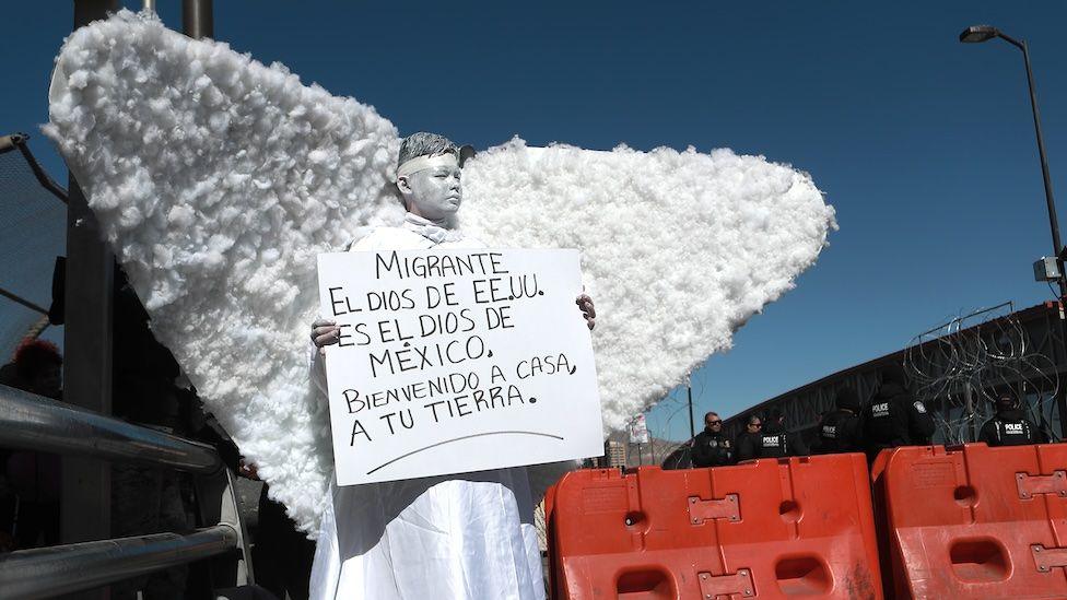 A member of a religious group dressed as an angel takes part in a demonstration at the Ciudad Juarez international crossing from Mexico to the US. They hold up a sign written in Spanish protesting against the the immigration policies of Donald Trump. It reads "Migrants, the God in the US is the God in Mexico. Welcome back home to your land." 