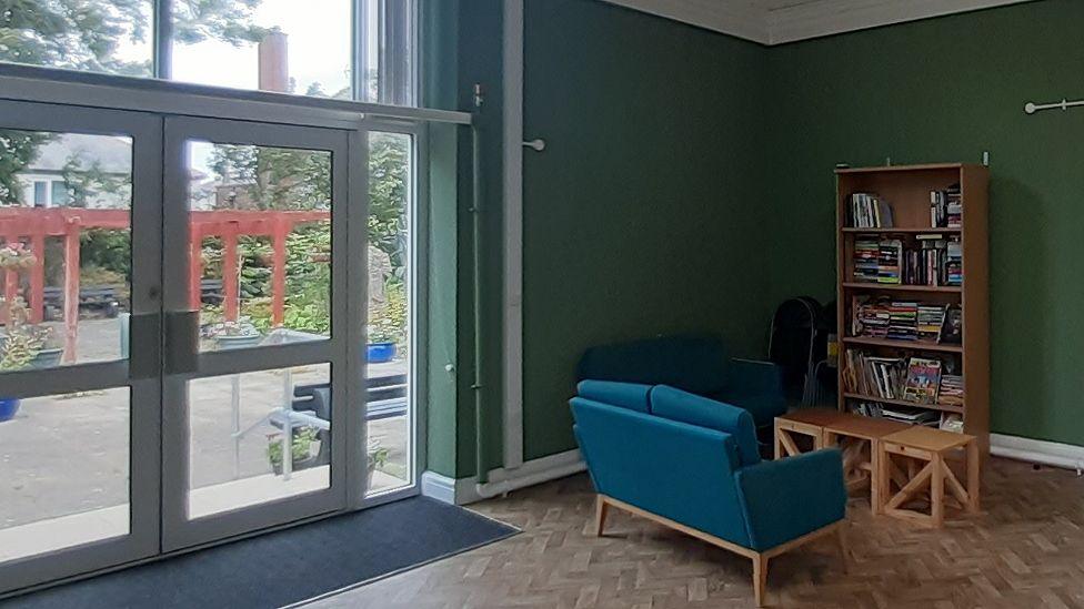 A room with green painted walls showing a sofa and bookcase creating a reading corner, and large patio doors opening out on to a garden with a large wooden pergola