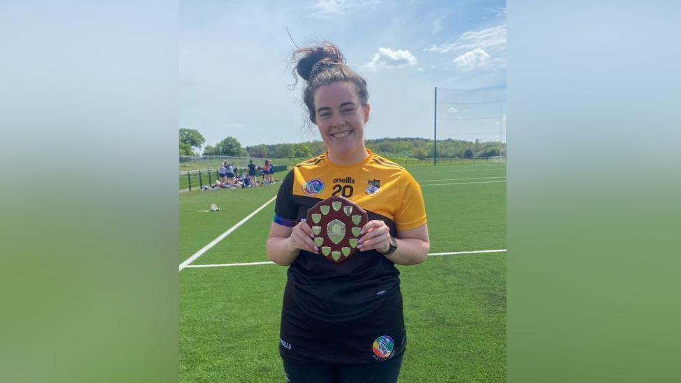 A woman in a yellow and black GAA jersey. She is holding a brown and silver trophy.