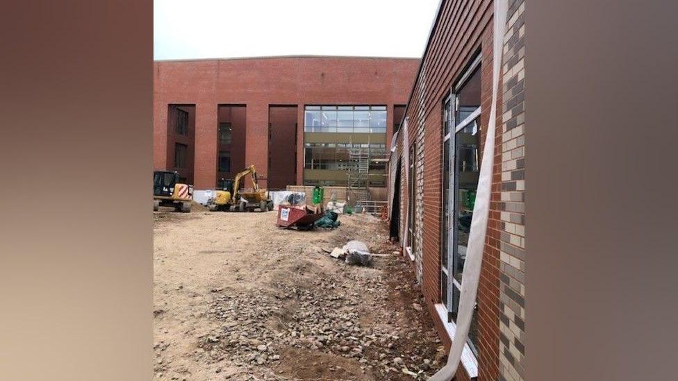 A red brick building with large windows has dirt on the ground in front of it, with a yellow digger, a skip and construction equipment.