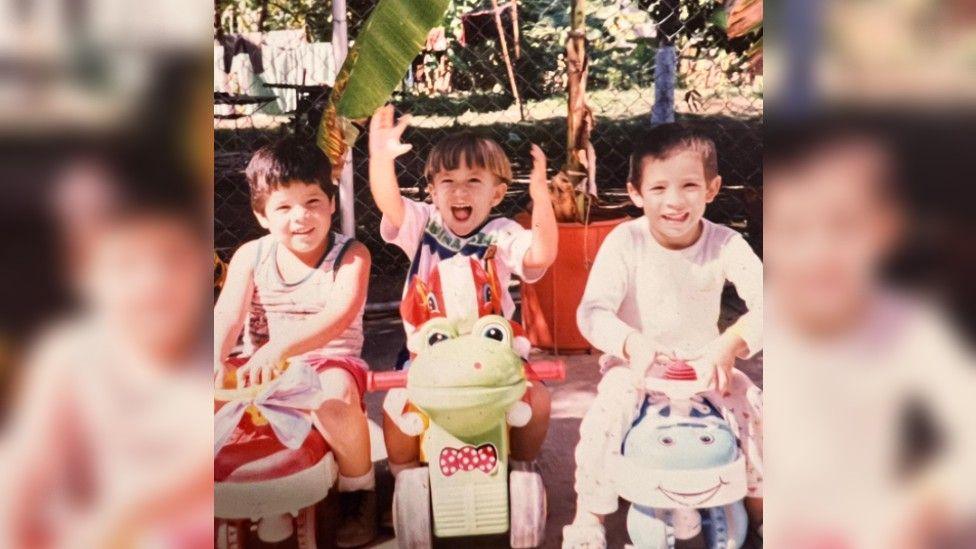 Ms Wailer as a young child on a toy bike with two cousins sitting on similar child toys.