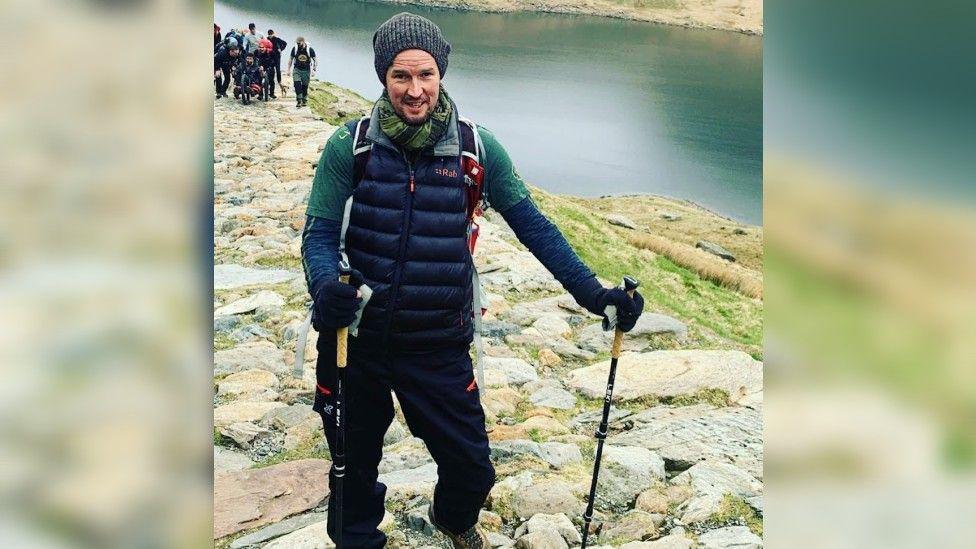 Jamie Rudd in walking gear by a river with a group of people in the background