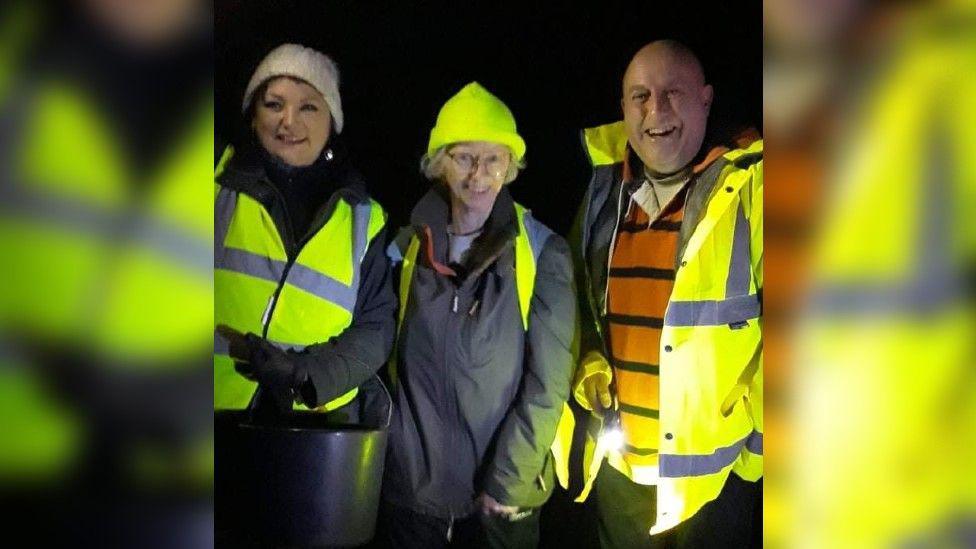 Three people at night in high-vis jackets smiling to camera, holding a bucket and a torch