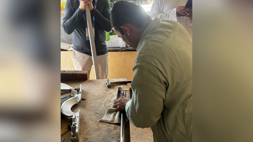 A carpenter fixes Matthew Selt's snooker cue. He is wearing a green shirt and holding the cue on a table, along with some tools.